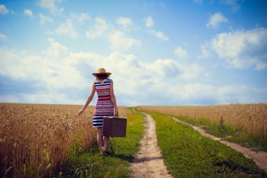Woman wearing hat with suitcase on road in wheat field