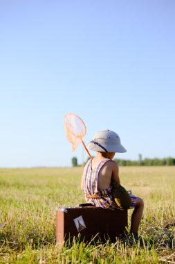Little boy with ring-net sitting on suitcase in summer countryside over blue sky background