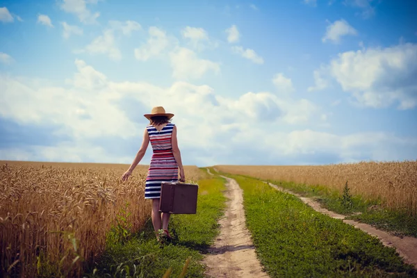 Woman wearing hat with suitcase on road in wheat field