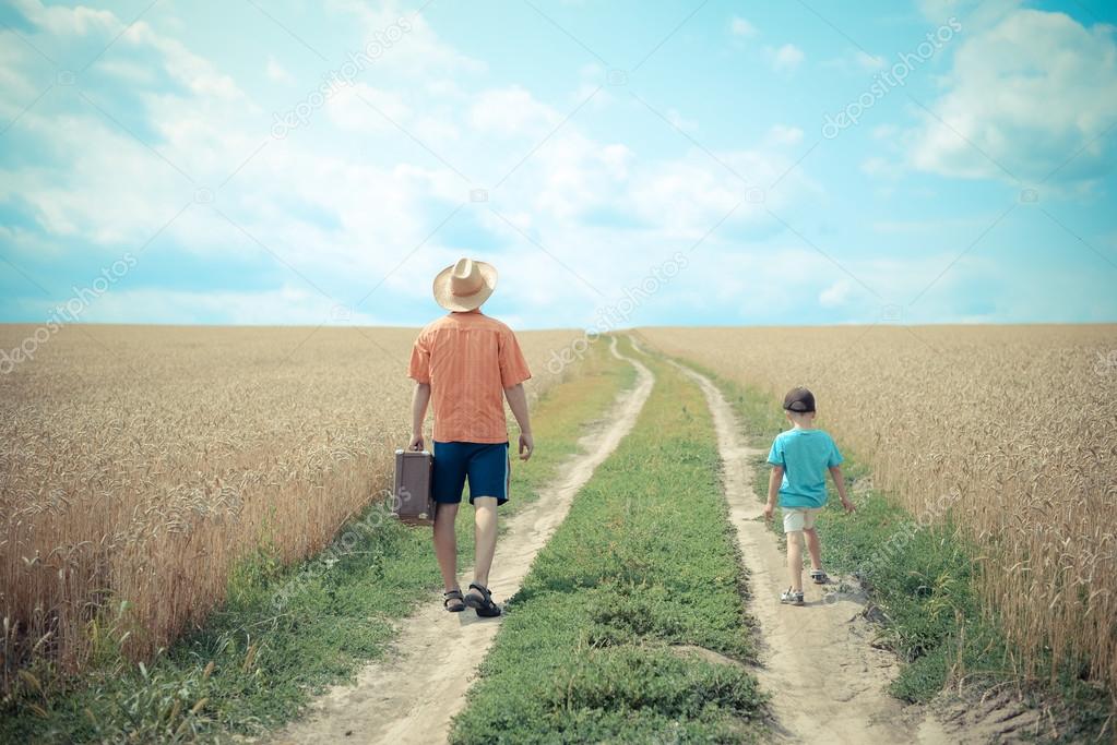 Man with valize and boy walking on road between field of wheat