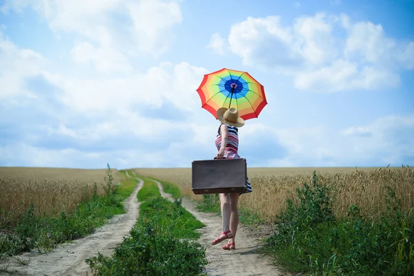 Mulher bonita com guarda-chuva arco-íris e mala velha na estrada rural — Fotografia de Stock