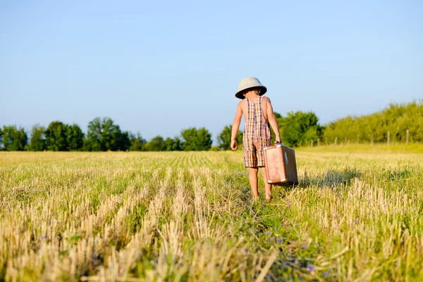 Little boy walking away carrying big old suitcase in field — 图库照片