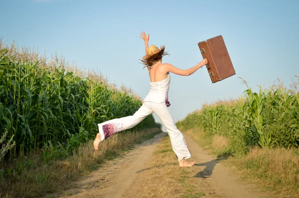 Young girl jumping with suitcase on road in corn field — Stock Photo, Image