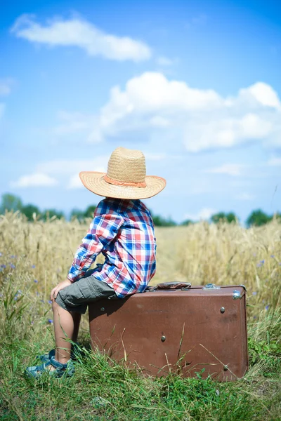 Little boy wearing cowboy hat sitting on suitcase in countryside — Stock Fotó