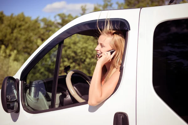 Picture of blond girl speaking phone and looking from car window. Young woman excited and smiling while talking with someone on sunny countryside — Stock Photo, Image