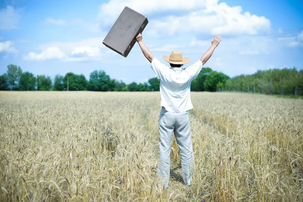 Happy man wearing hat with suitcase rising hands in field — Stock fotografie