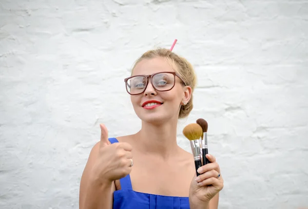 Блондинка в хипстерских очках лижет кисточками для макияжа улыбаясь — стоковое фото