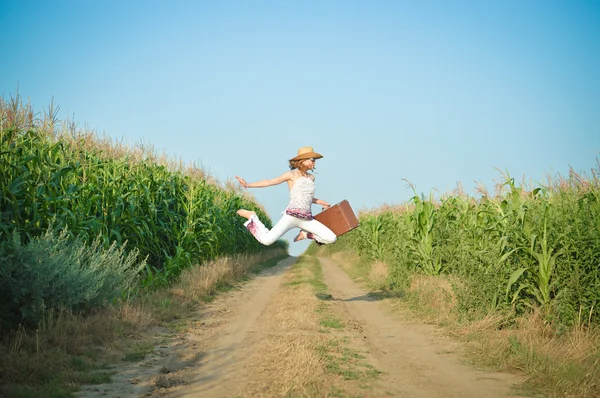 Young girl jumping with suitcase on road in corn field — Stock Photo, Image