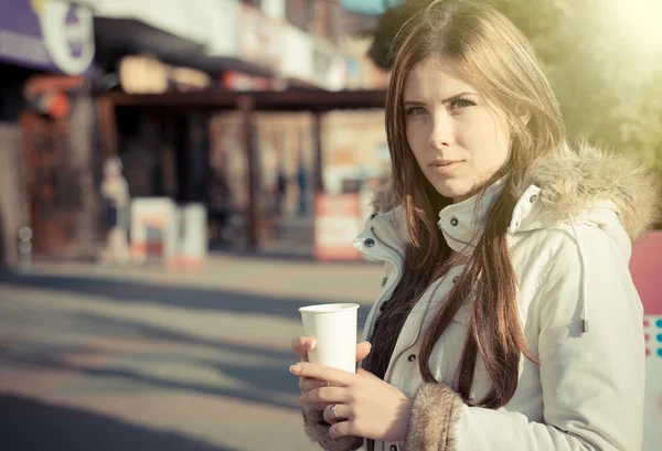 Pretty young woman in coat holding paper cup on street — ストック写真