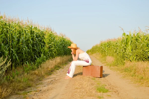 Mulher usando chapéu sentado na mala no meio do campo — Fotografia de Stock