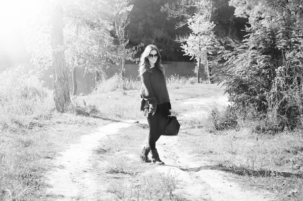 Picture of young pretty lady in sunglasses with bag walking on autumn countryside road and enjoying sunshine on sunny outdoors — Stock fotografie