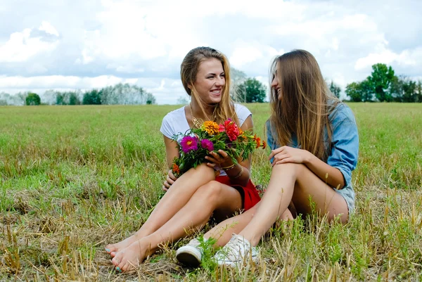 Two charming girls sitting on grass with wildflowers and talking — Stockfoto