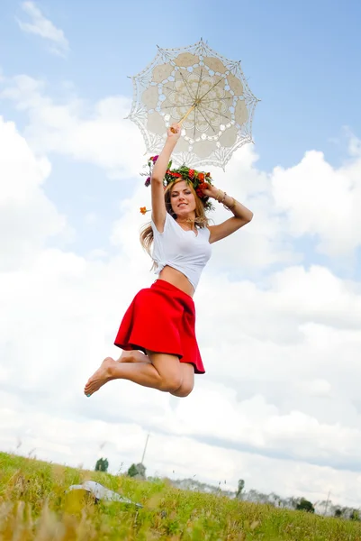 Радостная девушка в цветочном венке с кружевным зонтиком прыгает по лугу — стоковое фото