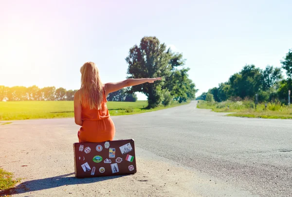Young woman sitting on suitcase and hitchhiking car in countryside — ストック写真