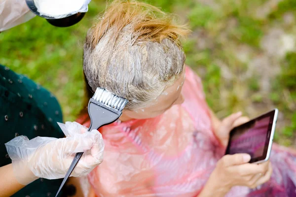 Peluquería para colorear el cabello con cepillo grande mientras el cliente lee el teléfono inteligente — Foto de Stock