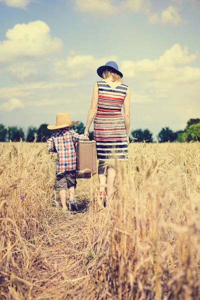 Mother and son walking along wheat field and carrying heavy suitcase — Stockfoto