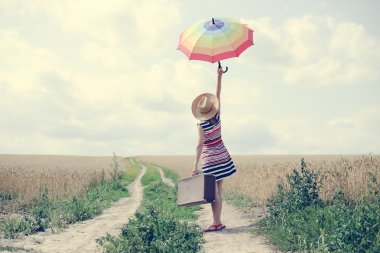 Woman with suitcase and umbrella standing on road between wheat over blue sky background