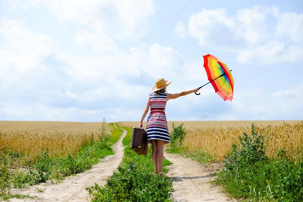 Girl holding suitcase and umbrella standing on road between wheat