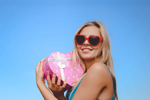 Bella ragazza in occhiali da sole fantasia in possesso di scatola regalo a forma di cuore rosa — Foto Stock