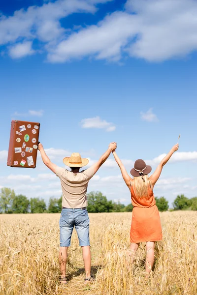 Happy exciting couple with valise rising hands together in wheat field — Stock Photo, Image