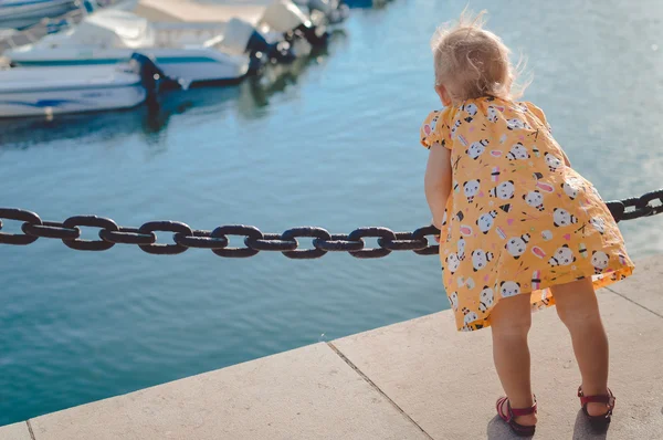 Little baby girl standing on embankment beside deep blue water — Stock Photo, Image