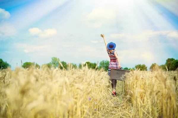 Beautiful young lady wearing hat with suitcase in wheat field — Stock Photo, Image