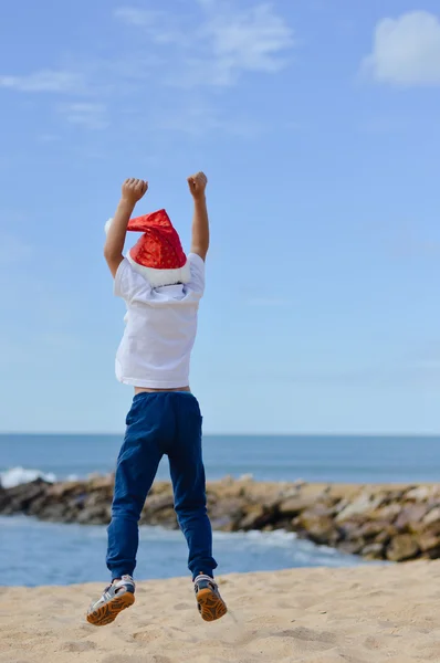 Backview of joyful little boy in Santa hat jumping on seashore — стокове фото