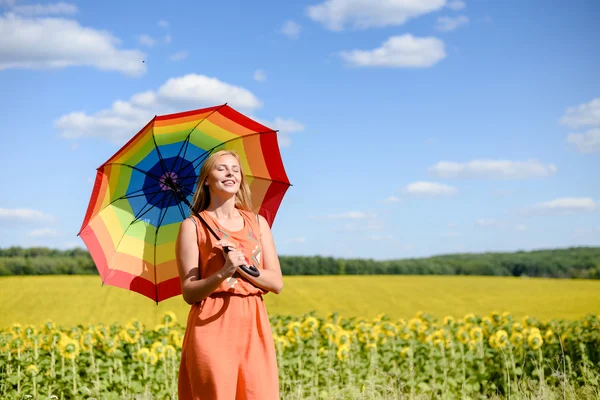 Joyful beautiful young lady with rainbow umbrella standing beside sunflowers — Stock Photo, Image