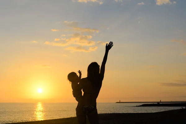 Silhouette of young exciting woman with hand up holding baby on dramatic seascape background — Stock Photo, Image