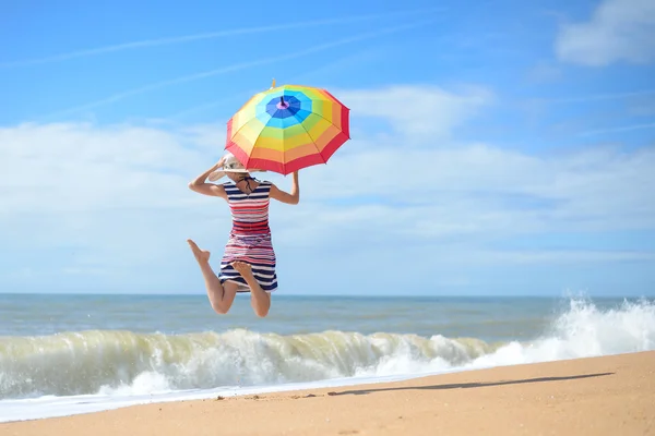 Schöne junge Frau mit Regenbogenschirm springt an sonnigem Meeresufer — Stockfoto