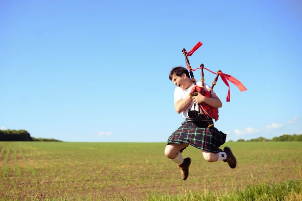 Picture of male jumping high with pipes in Scottish traditional kilt on green outdoors copy space summer field background — Zdjęcie stockowe