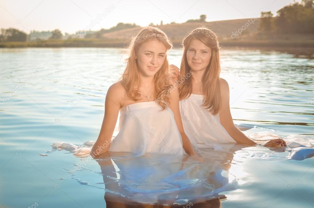 Portrait of two beautiful princess young ladies in white dresses on summer sunset water outdoors background