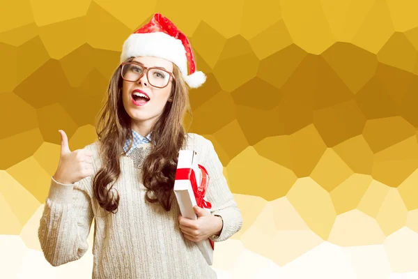 Portrait of showing thumb up and holding present box beautiful young lady in Christmas hat, glasses happy smiling and looking at camera — Zdjęcie stockowe