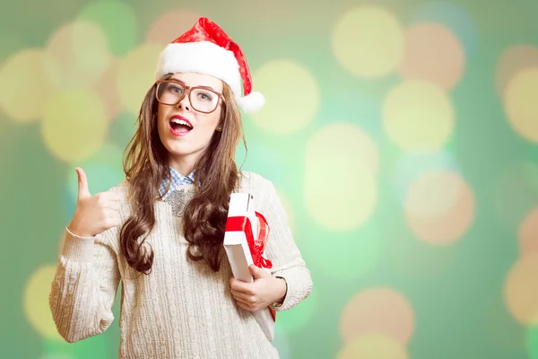 Showing thumb up and holding present box beautiful young lady in Christmas hat, glasses happy smiling and looking at camera — Stock Photo, Image