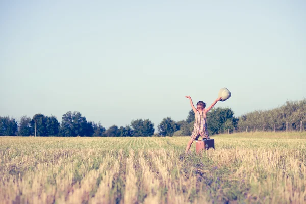 Happy boy with raised hands and suitcase in country field — Stok fotoğraf