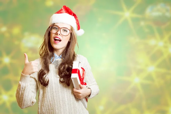 Showing thumb up and holding present box beautiful young lady in Christmas hat, glasses happy smiling and looking at camera — 图库照片