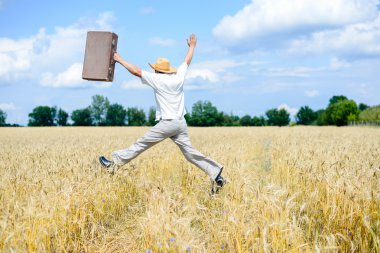 Male in hat jumping with retro suitcase on wheat field