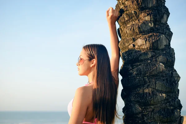 Picture of beautiful young woman in pink bikini and sunglasses standing beside palm tree. Sideview of pretty girl smiling on summer blue sky background. — Stock Photo, Image