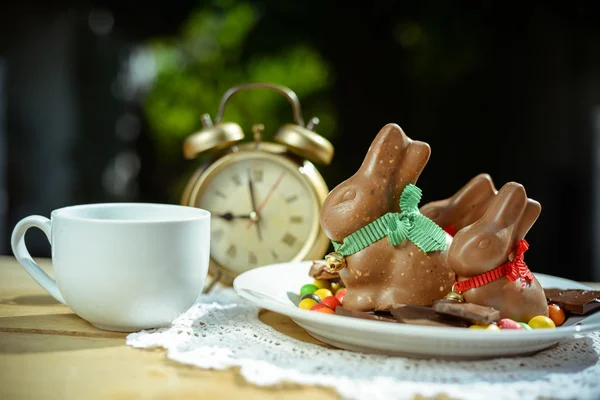 Plate with chocolate bunnies and candies beside retro alarm clock — Stock Photo, Image