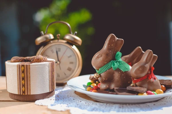 Plate with chocolate bunnies beside retro alarm clock and giftbox — Stock Photo, Image