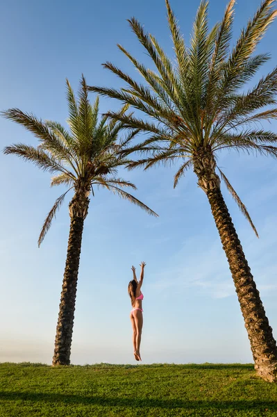 Picture of beautiful young excited lady in pink bikini dancing beside palm tree. Pretty girl flying or jumping on summer blue sky background. — Stock Fotó