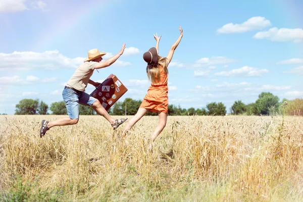 Jovem casal alegre se divertindo no campo de trigo. Homem e mulher excitados correndo com mala de couro retro no céu azul ao ar livre — Fotografia de Stock