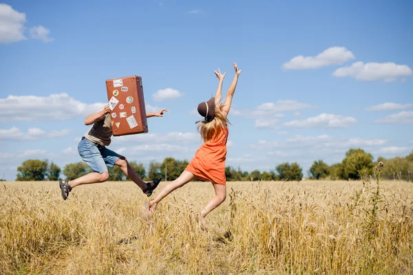 Fröhliches junges Paar, das Spaß im Weizenfeld hat. Aufgeregter Mann und Frau läuft mit Retro-Lederkoffer auf blauem Himmel — Stockfoto