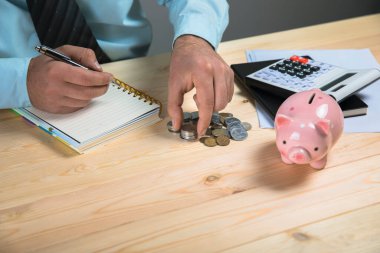 Hesaplamalar yapar ve bir deftere yazar. Yanında da kumbara ve bozukluklar vardır.