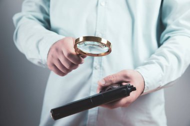 Genç bir adam elinde silah tutuyor ve büyüteçle bakıyor.