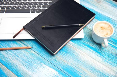 Masada dizüstü bilgisayar, not defteri, kalem ve kahve var.