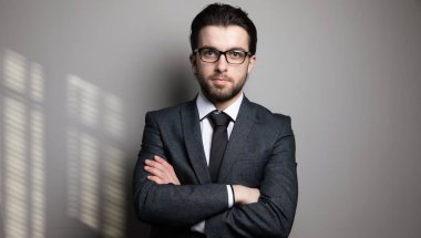 Takım elbiseli ve gözlüklü bir adam gri bir arka planda kollarını kavuşturmuş duruyor.