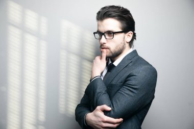 Takım elbiseli ve gözlüklü bir adam gri arka planda çenesine dokunur ve düşünür.