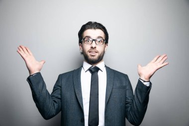 Takım elbiseli ve gözlüklü bir adam gri arka planda şaşkın şaşkın duruyor.