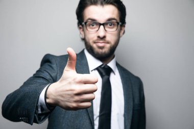 Takım elbiseli ve gözlüklü adam gri arka planda baş parmağını gösteriyor.